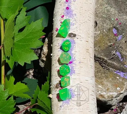 Smeraldo Impuro 2 Set di dadi Cloudy Emerald: sette dadi poliedrici verdi traslucidi con numeri gialli in fila su un tronco di betulla