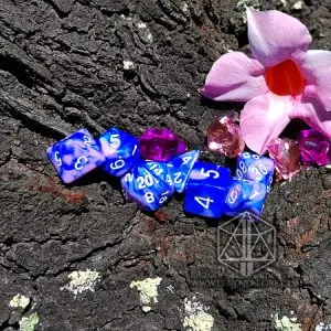 Set di dadi GdR Evening: dadi poliedrici blu e lilla marmorizzati con numeri bianchi su corteccia, accanto a un fiore rosa
