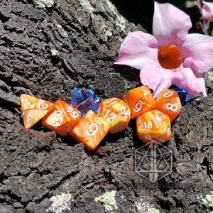 Set di dadi GDR Flames: sette dadi poliedrici marmorizzati arancio e giallo con numeri bianchi su corteccia accanto a un fiore rosa