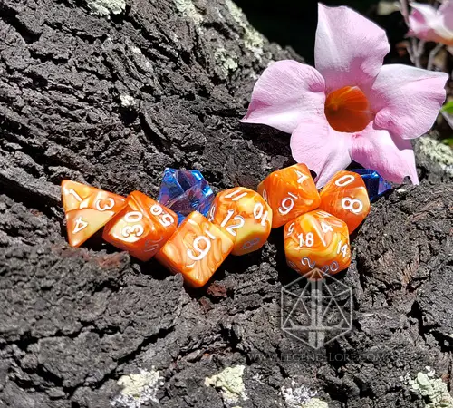 Flames RPG dice set: seven swirled orange and yellow polyhedral dice with white numbers on tree bark beside a pink flower