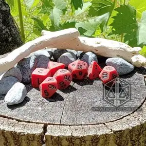 Set di dadi GdR Hardened Rust: sette dadi poliedrici rossi opachi effetto antico con numeri neri su un ceppo tra sassi
