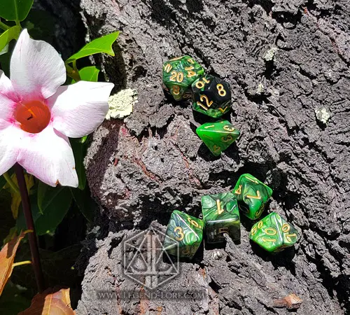 Jungle RPG dice set: seven green and black swirled polyhedral dice with gold numbers on tree bark beside a pink flower
