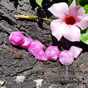 Set di dadi GDR Roses and Flowers: sette dadi poliedrici rosa e bianchi marmorizzati su corteccia accanto a un fiore rosa