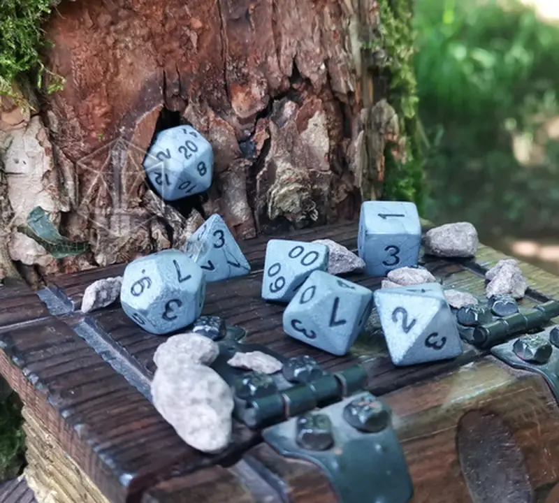 Set di dadi GDR Rough Stone: sette dadi poliedrici grigi effetto pietra con numeri neri su un baule di legno vicino a un tronco