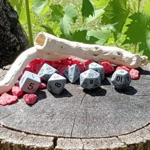 Set Rough Stone di sette dadi poliedrici grigi opachi con numeri neri su un ceppo con sassi rossi e legno levigato