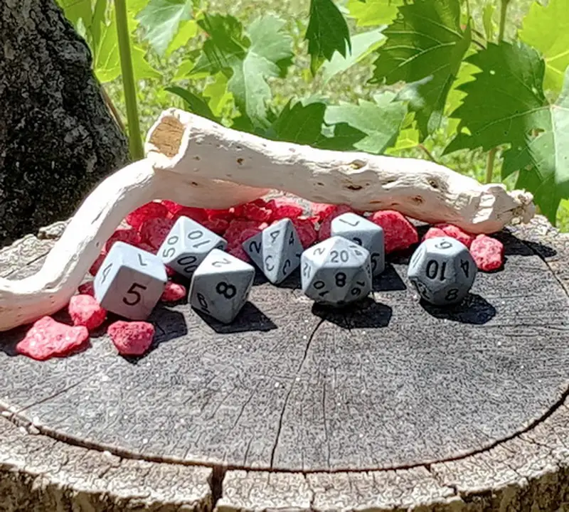 Set Rough Stone di sette dadi poliedrici grigi opachi con numeri neri su un ceppo con sassi rossi e legno levigato
