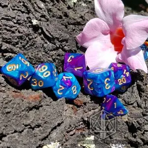 Set di dadi GDR Vitriol: sette dadi poliedrici blu e viola marmorizzati con numeri dorati su corteccia accanto a un fiore rosa