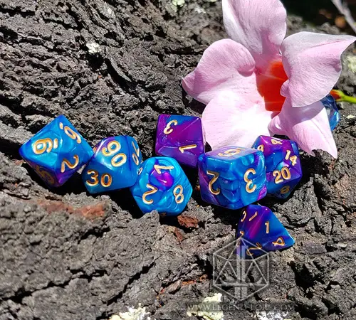 Vitriol RPG dice set of seven blue and purple swirled polyhedral dice with gold numbers on bark beside a pink flower