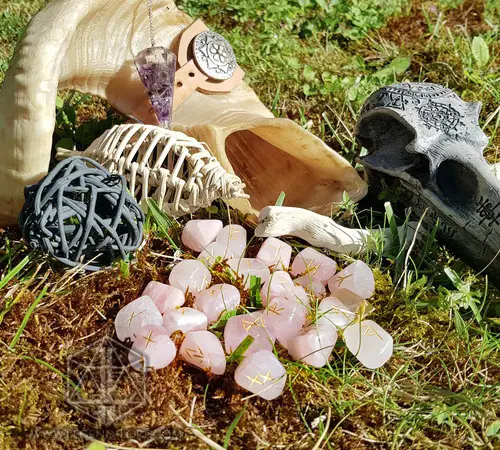 Rune divinatorie Crystal Petals in quarzo rosa con simboli dorati sul muschio, accanto a corno, teschio intagliato e pendolo