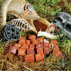 Rune divinatorie Cursed Spines in diaspro rosso con simboli dorati sul muschio, accanto a un corno d'ariete e un teschio