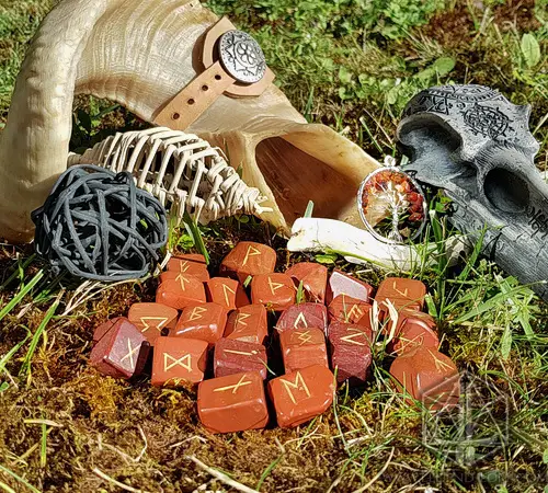 Cursed Spines Divination Runes: red jasper stones with gold Elder Futhark symbols on moss beside a ram horn and skull