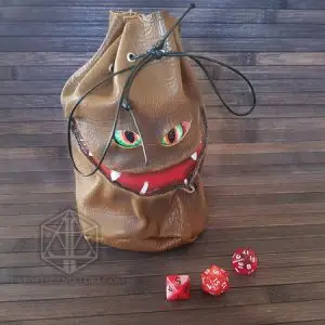 RPG ACCESSORIES - EN 5 Evil Bag brown leather dice bag with green eyes, red grinning mouth and black drawstring, beside three red dice