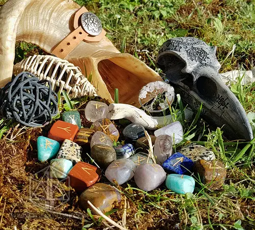 Iridescent Cornucopia divination runes: mixed gemstones engraved with gold runes, beside a horn and raven skull on grass
