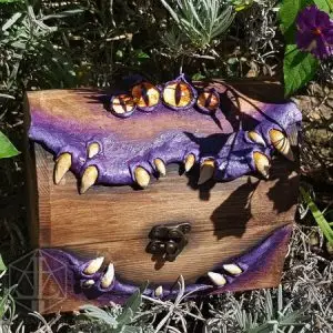 RPG ACCESSORIES - EN 3 Mimic Lord wooden dice box with purple sculpted gums, cream fangs, four orange eyes and a brass clasp, set among plants