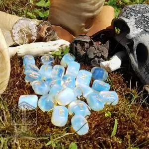 Rune divinatorie Moon Tears in opalite traslucida con simboli dorati del Futhark su muschio, accanto a un teschio intagliato
