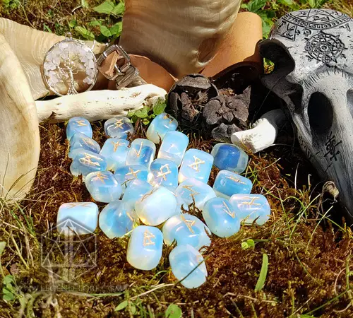 Moon Tears divination runes, translucent opalite stones with gold Elder Futhark symbols on moss beside a carved skull
