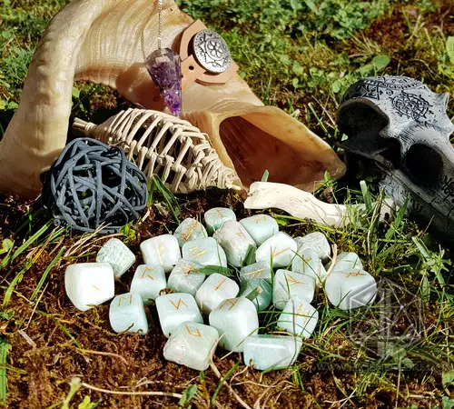 Rune divinatorie Wind Soul in amazzonite verde chiaro con simboli dorati sull'erba, accanto a corno, pendolo e teschio
