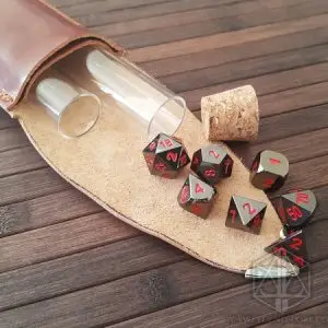 Set Blood Pact di sette dadi poliedrici in metallo nero con numeri rossi, accanto a custodia in pelle, fiale e tappo di sughero