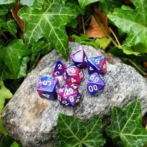 Set di dadi GdR Cruelty: sette dadi poliedrici blu e viola sfumati con numeri bianchi su una roccia grigia tra foglie d'edera