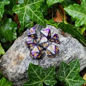 Set di dadi Necromancer: sette dadi poliedrici grigi e viola marmorizzati con numeri dorati su una roccia tra foglie d'edera
