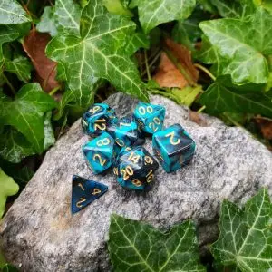 Set di sette dadi poliedrici Sea Serpent in turchese e nero marmorizzato con numeri dorati su una roccia tra foglie d'edera