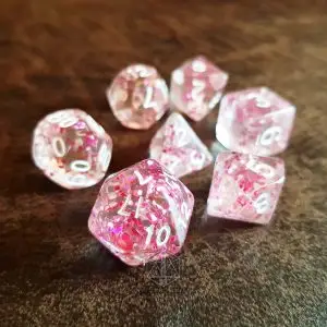 Set Raining Roses di sette dadi poliedrici trasparenti con inclusioni rosa e numeri bianchi su superficie marrone
