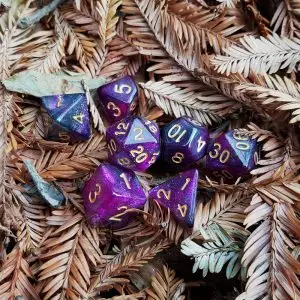 Set Sparkling Velvet di sette dadi poliedrici viola glitterati con numeri dorati su aghi di conifera secchi