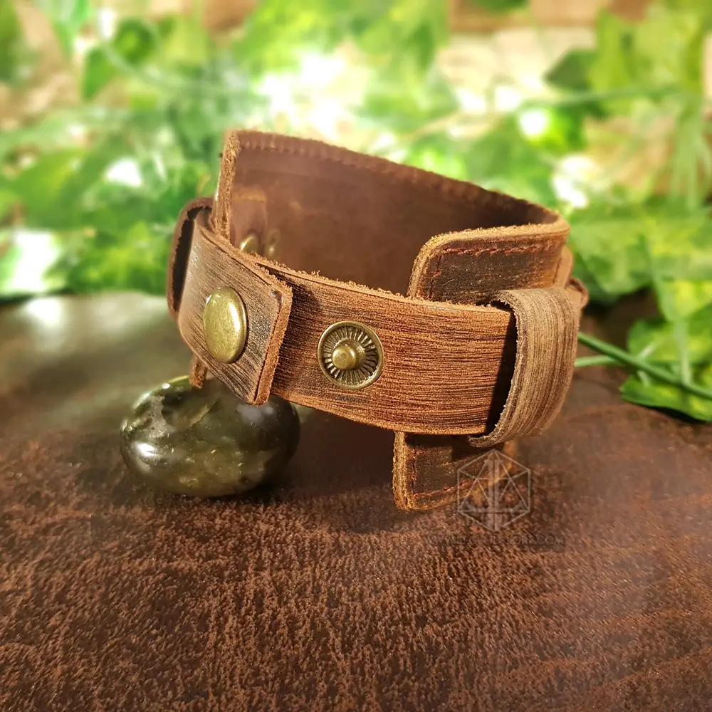 Ancient Oath Viking bracelet in distressed brown leather with brass snap buttons, beside a dark green stone and leaves