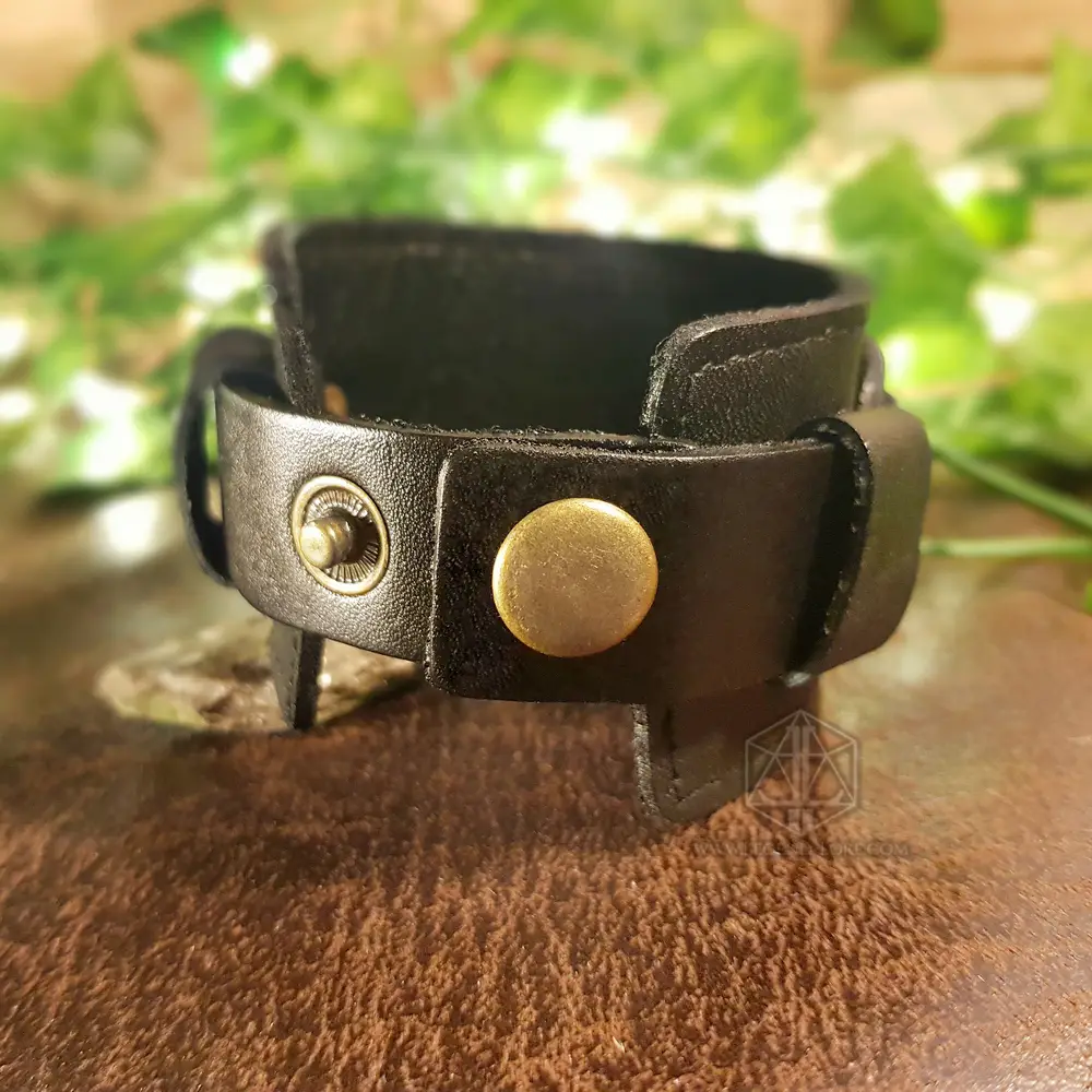 Bracciale vichingo Dark Oath in pelle nera con bottone a pressione in ottone, su pelle scamosciata marrone tra foglie verdi
