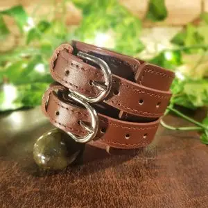 Bracciale vichingo The Wild Big Wheel in pelle marrone con due cinturini e fibbie argentate, accanto a una pietra verde