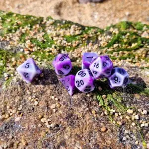 Set Toxic Coral di sette dadi poliedrici bianchi e viola marmorizzati con numeri neri su una roccia con muschio e sabbia