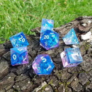 Set di sette dadi in resina a spigolo vivo Aurora Shardglass, blu e viola con scaglie iridescenti, su corteccia vicino all'erba