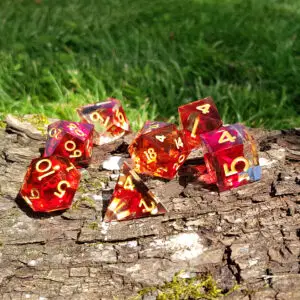 Set di sette dadi in resina a spigolo vivo Phoenix Shards, rosso traslucido con numeri dorati, su corteccia vicino all'erba