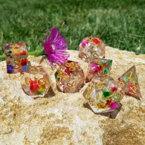 Set di sette dadi Wildflower Blossoms in resina trasparente a spigoli vivi con fiori secchi e numeri dorati su una roccia