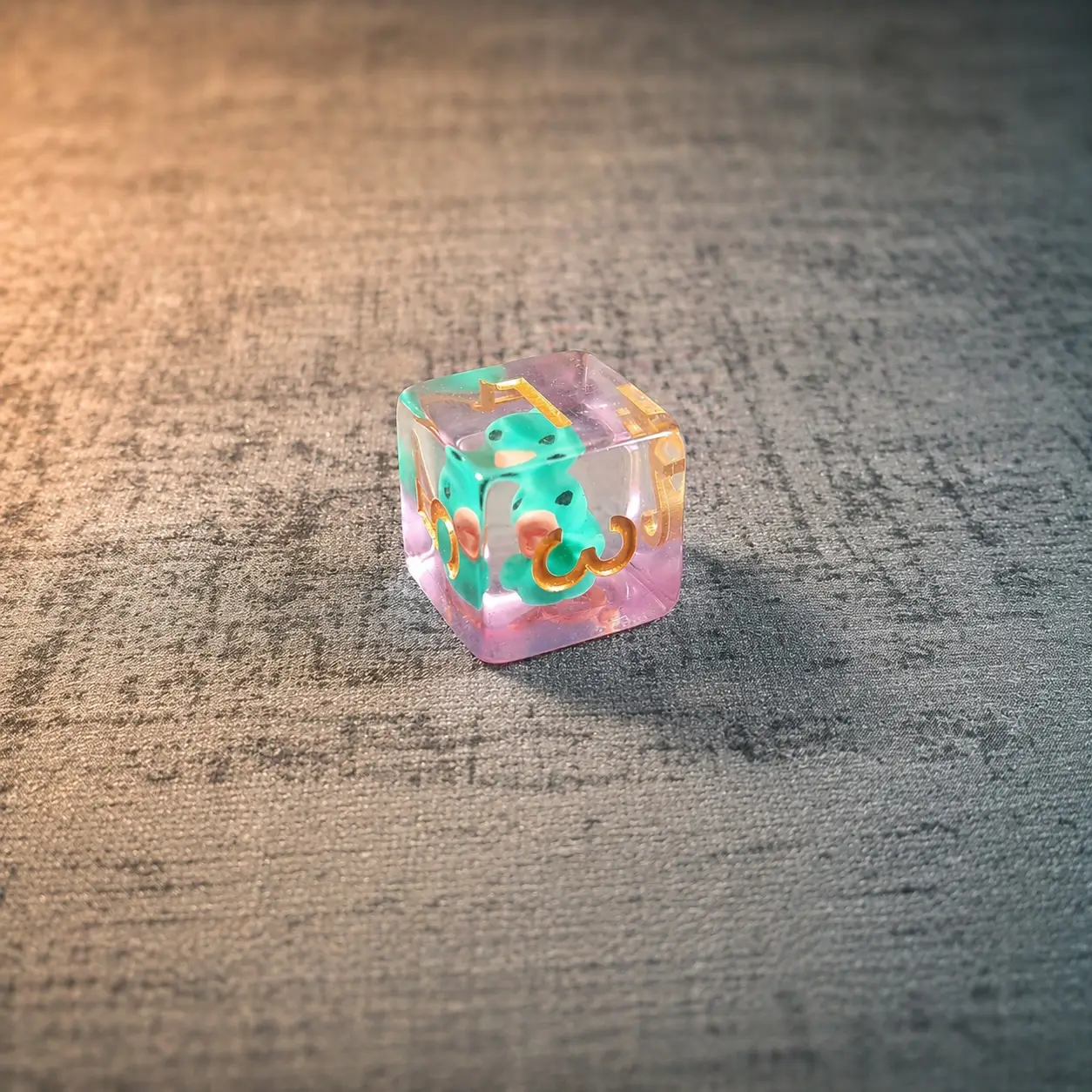 Single D6 from the PERRY the Duck dice set, clear pink resin with a teal duck inside and gold numbers, on grey fabric